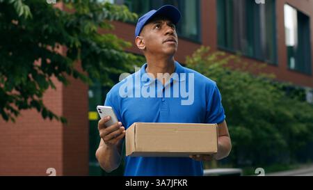 Afroamerikanischer Kuriermann verwirrter Blick auf Handy GPS Karte online Suche Standort Adresse Suche um Wartebox Zustellung Postarbeiter Stockfoto