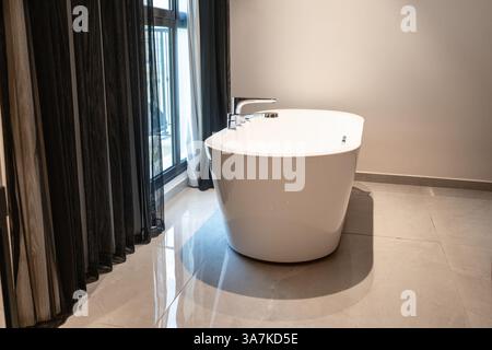 Moderne weiße freistehende Badewanne in einem stilvollen Badezimmer mit großem Fenster, schwarzen Vorhängen und poliertem Steinboden, die an Luxus und modernen Komfort erinnern. Stockfoto