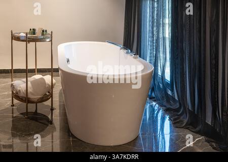 Stilvolle freistehende Badewanne mit gerollten Handtüchern und Spa-Produkten auf einem Beistelltisch in einem ruhigen, sanft beleuchteten Luxusbad mit schwarzem Marmorboden und transparenten Vorhängen. Stockfoto