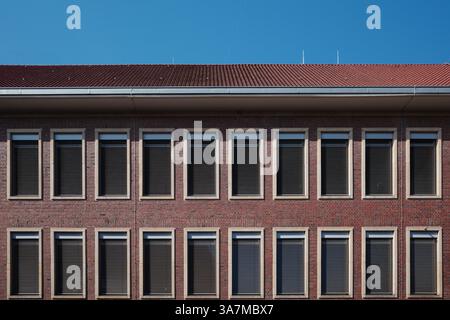 Ein rotes Backsteingebäude mit einem symmetrischen Gitter aus rechteckigen Fenstern, die jeweils mit geschlossenen horizontalen Jalousien ausgestattet sind. Stockfoto