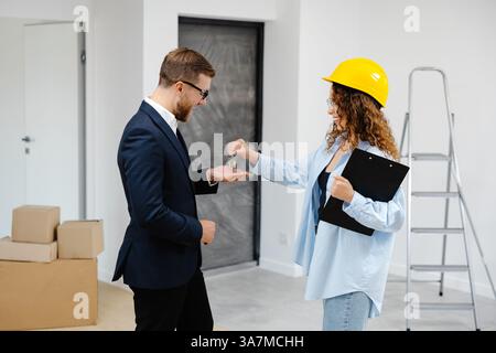 Designerin im Helm mit Dokumenten, die Schlüssel von der Wohnung an einen jungen Geschäftsmann geben, während er im Zimmer steht. Stockfoto