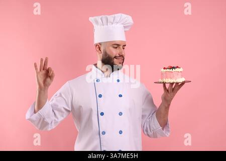 Glücklicher Konditor in Uniform, der Kuchen mit Beeren hält und eine gute Geste auf rosafarbenem Hintergrund zeigt Stockfoto