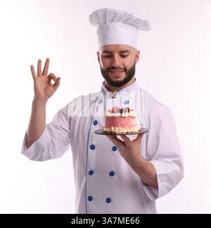 Glücklicher Konditor in Uniform, der leckeren Kuchen mit Beeren hält und eine gute Geste auf weißem Hintergrund zeigt Stockfoto