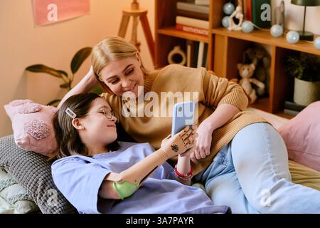 Die blonde Mutter und ihre brünette Tochter mit Prothesen sitzen auf dem Sofa, und einer hält das Smartphone, während er lächelt Stockfoto