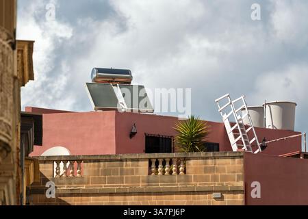 Sonnenkollektoren auf dem Dach eines Hauses vor einem stürmischen Himmel. In der Nähe sind zusätzliche Anlagen, eine Leiter und Wassertanks aus Metall installiert. Stockfoto