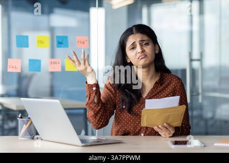 Porträt einer verärgerten und enttäuschten Frau am Arbeitsplatz im Büro. Büroangestellter, der in die Kamera blickt, unglücklich Briefumschlag hält, mit schlechten Nachrichten in der Hand. Stockfoto