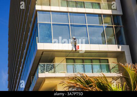 Fensterreiniger mit rotem Eimer, der an einem Glashochhaus vor blauem Himmel im tropischen Resort arbeitet. Mexiko. Cancun. Stockfoto