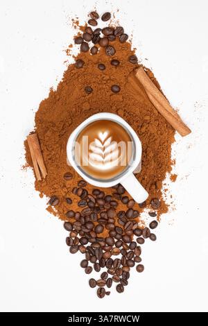 Eine Tasse Cappuccino mit Latte Art umgeben von Kaffeebohnen, Zimtstangen und gemahlenem Kaffee auf weißem Hintergrund. Stockfoto