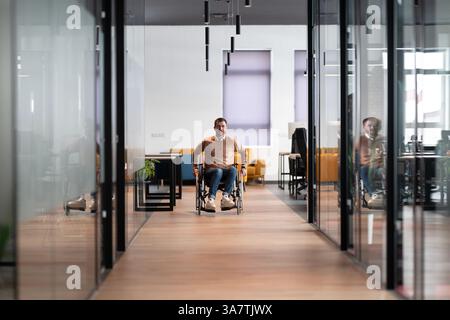 Moderner Startup Coworking-Büroflur mit Glaswänden, mit einem Mann im Rollstuhl. Der Raum bietet einen integrativen, zugänglichen Arbeitsplatz Stockfoto