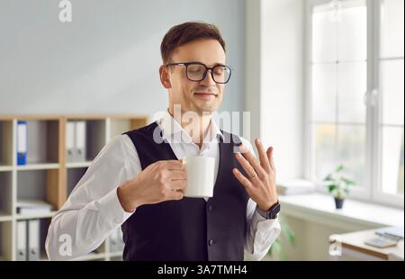 Ein glücklicher junger Geschäftsmann genießt morgens den Geruch seines Kaffees bei der Arbeit Stockfoto