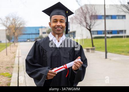 Lächelnder afroamerikanischer Absolvent mit Abschlusskleid und Mütze, der ein Diplom hält, auf dem Universitätscampus spazieren geht Stockfoto