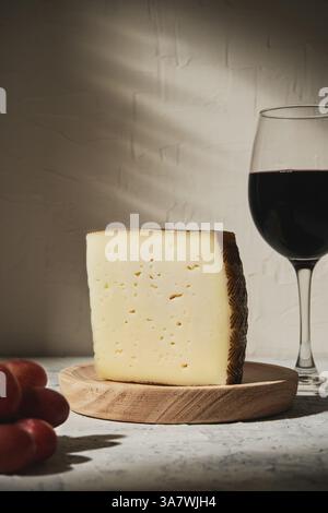 Dreieck Stück köstlichen frischen Hartkäse auf Holz platziert Board in der Nähe Trauben und Rotwein auf dem Tisch in Licht studio Stockfoto