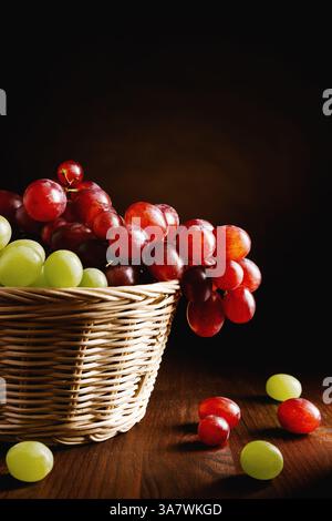 Weidenkorb gefüllt mit appetitlich frischen grünen und roten Trauben Auf Holztisch gegen schwarzen Hintergrund platziert Stockfoto