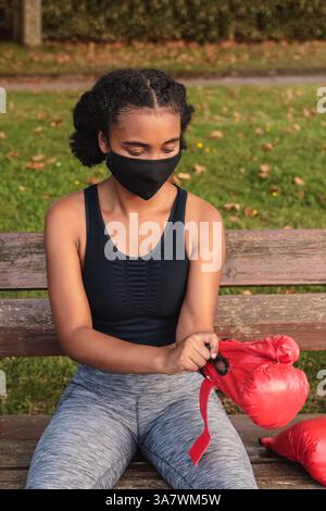 Von oben anonymen jungen schwarzen weiblichen Athleten in Schutz Maske und Sportbekleidung tragen rote Boxhandschuhe, während sie sitzen Bank im Park vor w Stockfoto