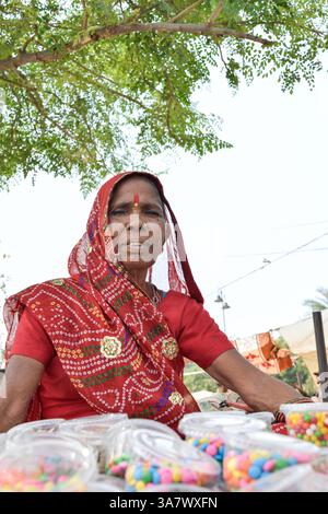 Ältere Frau in rotem Saree, die auf dem Straßenmarkt in Jaipur, Indien, Lebensmittel und Süßigkeiten verkauft. Stockfoto