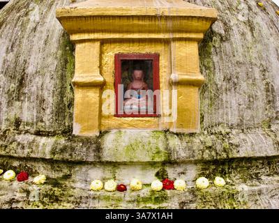 Kleine Buddha-Statue in einer Nische auf der moosgestreiften Kuppel von Boudhanath Stupa, eingerahmt von Ringelblumen- und Chrysanthemen-Opfergaben. Stockfoto