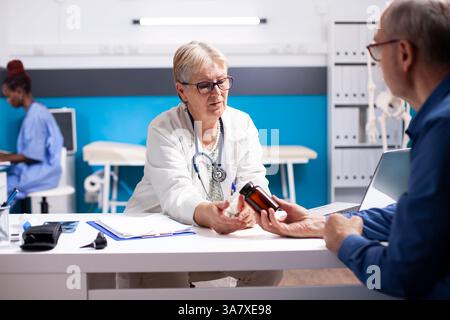 Älterer weißer Mann, der eine Medikamentenflasche hält, erhält Rat von einem älteren weiblichen Arzt über seinen Gesundheitsplan. Der leitende Arzt sitzt neben den pensionierten Patienten und gibt verschriebene Ergänzungen. Stockfoto