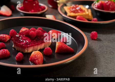 Vorderansicht eines Erdbeerdesserts mit Himbeeren auf einer schwarzen Keramikplatte im Vordergrund vor einem Hintergrund von Keksen mit Erdbeermarmelade und s Stockfoto