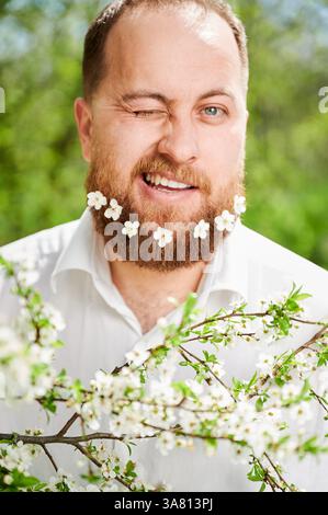 Mann-Allergie genießt nach Behandlung von Saisonallergie im Frühjahr. Porträt eines glücklichen Mannes mit kleinen weißen Blumen über seinem Bart, die im Frühling ein Auge vor dem Blütenbaum zuzwinkernd. Stockfoto