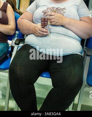Fettleibige weiße Frau, die in einem Wartezimmer des Krankenhauses saß und eine Tafel Schokolade hielt, mit ihrem Gesicht nicht sichtbar im Rahmen Stockfoto