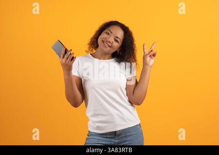 Fröhliche junge afroamerikanische Frau, die ein Smartphone hält und ein Friedenszeichen zeigt Stockfoto