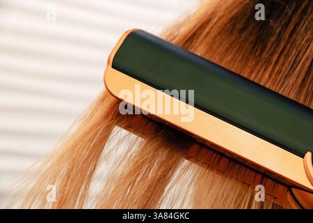 Detaillierte Aufnahme eines Haarglätters, der durch langes, hellbraunes Haar gleitet. Kosmetikmode und Styling-Werkzeuge Stockfoto
