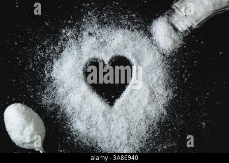 Herz aus Salz, das auf dem Tisch verstreut ist. In der Nähe befindet sich ein Metalllöffel mit Salz und ein offener Salzstreuer, mit dem Salz ausgegossen wird Stockfoto