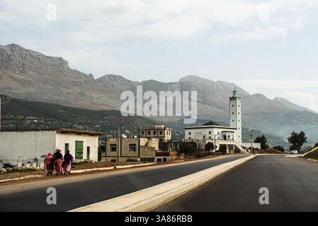 Ländliche Straße durch ein marokkanisches Dorf mit einer Moschee und hoch aufragenden Bergen in der Ferne, Marokko Stockfoto