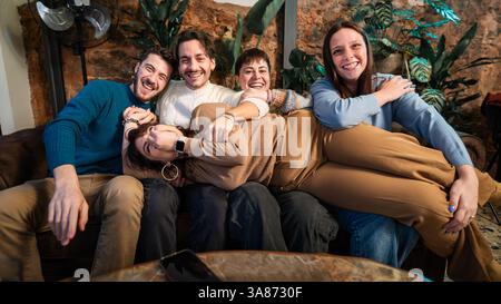 Junge, vielfältige Freunde halten ihren Freund über die Couch, lächeln und genießen ein fröhliches Indoor-Treffen mit spielerischer Energie. Stockfoto
