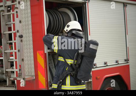 Ein Feuerwehrmann in voller Schutzausrüstung holt einen gewickelten Schlauch aus dem offenen Seitenfach eines roten Feuerwehrwagens, der einen Druckluftbehälter und einen Helm trägt Stockfoto