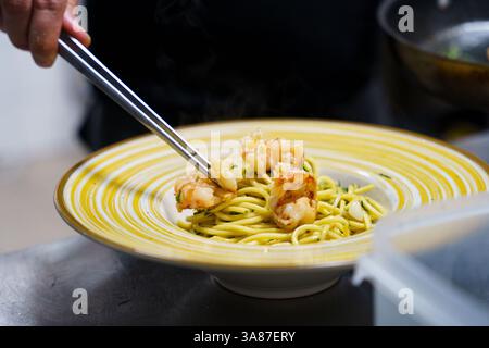 Ein Küchenchef legt gegrillte Garnelen vorsichtig mit einer Metallzange auf einen Teller Spaghetti aglio e olio. Die Pasta, die mit Olivenöl glänzt, wird garniert Stockfoto