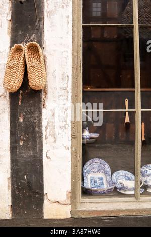 Rustikale Szene mit einem Paar gewebter Strohschuhe, die an einer verwitterten Wand neben einem antiken Schaufenster hängen, in dem Vintage-blau-weiße Porzellan zu sehen sind Stockfoto