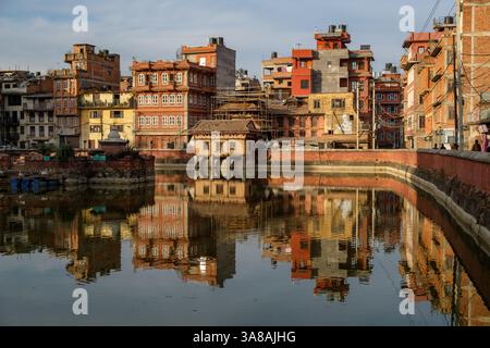 Gebäude spiegeln sich in Pimbahal Pokhari, Patan, Nepal. Die Menschen spazieren am historischen Teich vorbei, der eine Mischung aus traditioneller und moderner Architektur bietet. Stockfoto