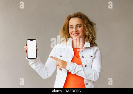 Frau mittleren Alters in einem orangefarbenen Oberteil und einer weißen Jacke, die ein Smartphone mit leerem Bildschirm hält und darauf zeigt, um mobile Apps oder Inhalte mit Co zu bewerben Stockfoto