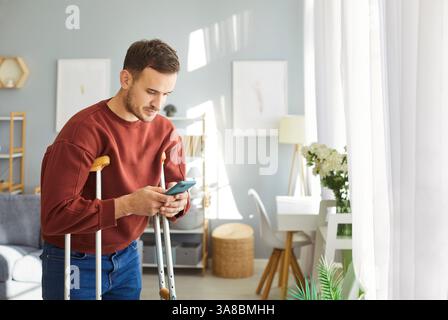 Junger behinderter Mann, der zu Hause am Fenster steht, mit Krücken, die das Handy in der Hand halten. Stockfoto