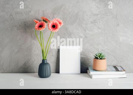 Rosafarbene Gerbera-Blüten in Vase mit leerem Bilderrahmen und Sukkulenten auf dem Tisch. Modell. Stockfoto