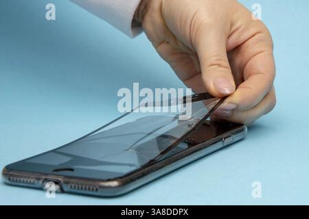 Weibliche Hände lösen das zerbrochene Schutzglas eines Smartphones auf blauem Hintergrund, Nahaufnahme, selektiver Fokus. Konzept: Glasersatz, Telefon r Stockfoto