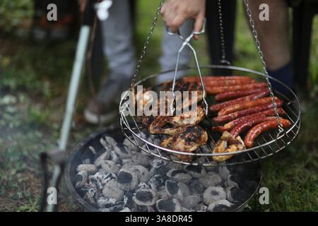 Sommer-Grill im Hinterhof mit Würstchen und Fleisch auf einem runden Grill – eine Hand reicht hinein, um die rauschende Szene zu verstärken. Stockfoto
