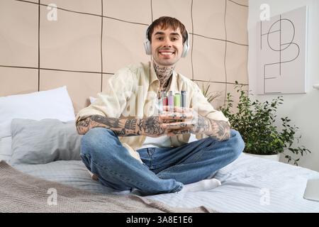 Junger gutaussehender Mann in Kopfhörern mit verschiedenen elektronischen Einweg-Zigaretten auf dem Bett zu Hause Stockfoto