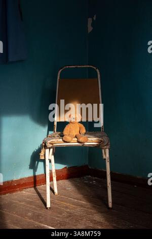 Brauner, weicher Teddybär, der auf einem Stuhl sitzt, in einem alten verlassenen Haus in der Ukraine, vergessenes, verlassenes Spielzeug Stockfoto