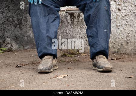 Schmutziger männlicher Maler in Arbeitsuniform in Farben sitzt auf einem Stuhl auf der Straße ruht, bricht, männliche Beine, harte Arbeit Stockfoto