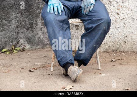 Schmutziger männlicher Maler in Arbeitsuniform in Farben sitzt auf einem Stuhl auf der Straße ruht, bricht, männliche Beine, harte Arbeit Stockfoto