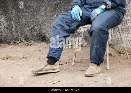 Schmutziger männlicher Maler in Arbeitsuniform in Farben sitzt auf einem Stuhl auf der Straße ruht, bricht, männliche Beine, harte Arbeit Stockfoto