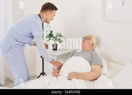 Junger, freundlicher Krankenpfleger, der einem Seniorpatienten mit iv-Tropfinfusion hilft. Stockfoto