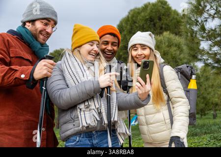 Vier glückliche Wanderer machen ein Selfie mit einem Smartphone, während sie die Wanderstöcke halten und einen Wintertag in einem Wald genießen Stockfoto