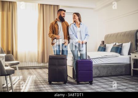 Bärtige Männer und Frauen mit braunen Haaren kamen im Hotel an. Pärchen stehen im Hotelzimmer mit ausgepackten Koffern. Stockfoto