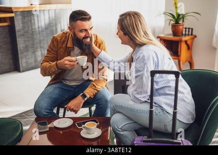 Fröhliches verheiratetes Paar kam im Hotel an. Sie sind im Urlaub, sitzen in der Lobby mit Koffern, trinken Kaffee und haben Spaß. Stockfoto