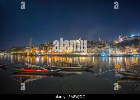 Porto, Portugal: Nachtblick auf den Fluss Douro mit beleuchteter Skyline und traditionellen Rabelo-Booten, die sich im Wasser spiegeln. Stockfoto