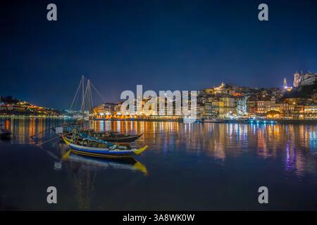 Porto, Portugal: Nachtblick auf den Fluss Douro mit beleuchteter Skyline und traditionellen Rabelo-Booten, die sich im Wasser spiegeln. Stockfoto