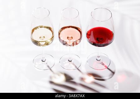Drei Weingläser gefüllt mit Rot-, weiß- und Rosenwein, anmutig auf einem hellen, minimalistischen Hintergrund. Eine lebendige Darstellung der Vielfalt der Weine Stockfoto